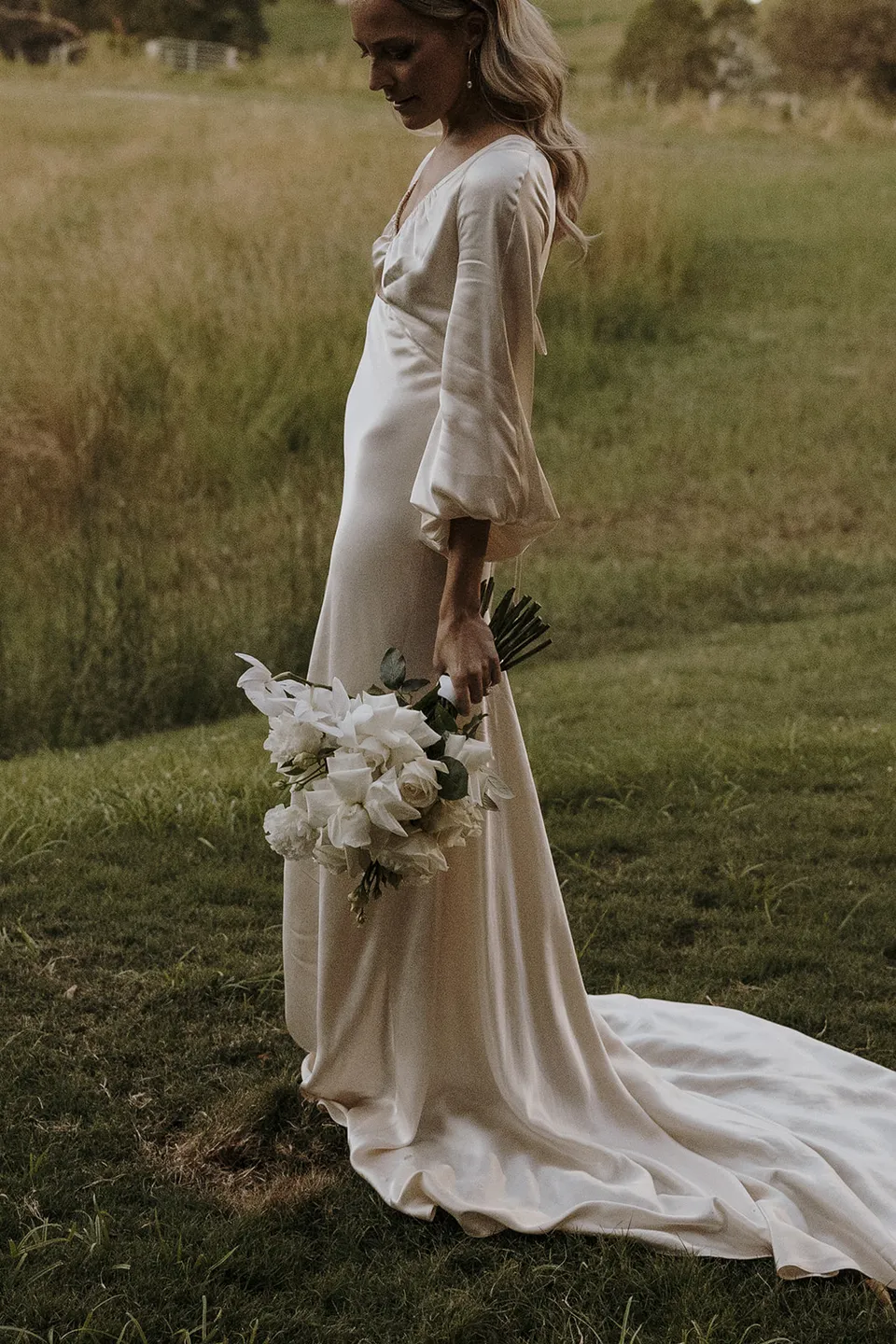 Gabrielle looking down at her white bouquet in the paddock, her ivory dress trailing through the grass at Roundhill Retreat in Obi Obi, Sunshine Coast Hinterland, for their Elopement Collective elopement.