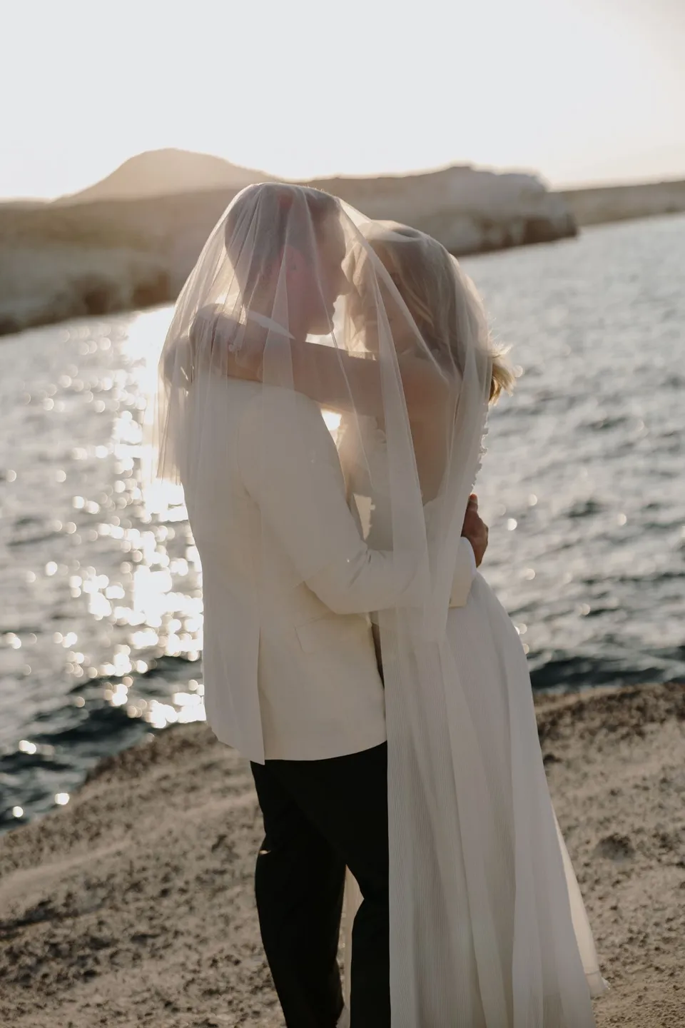 Georgina and Brad kissing in profile beside the sea, with her veil lifted by the breeze, at Sarakiniko on Milos in the Cyclades, Greece, during an Elopement Collective elopement. Photograph by George Bowden.
