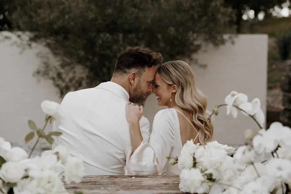 Gabrielle and Greg touching foreheads at a timber courtyard table surrounded by white flowers at Roundhill Retreat in Obi Obi, Sunshine Coast Hinterland, for their Elopement Collective elopement.