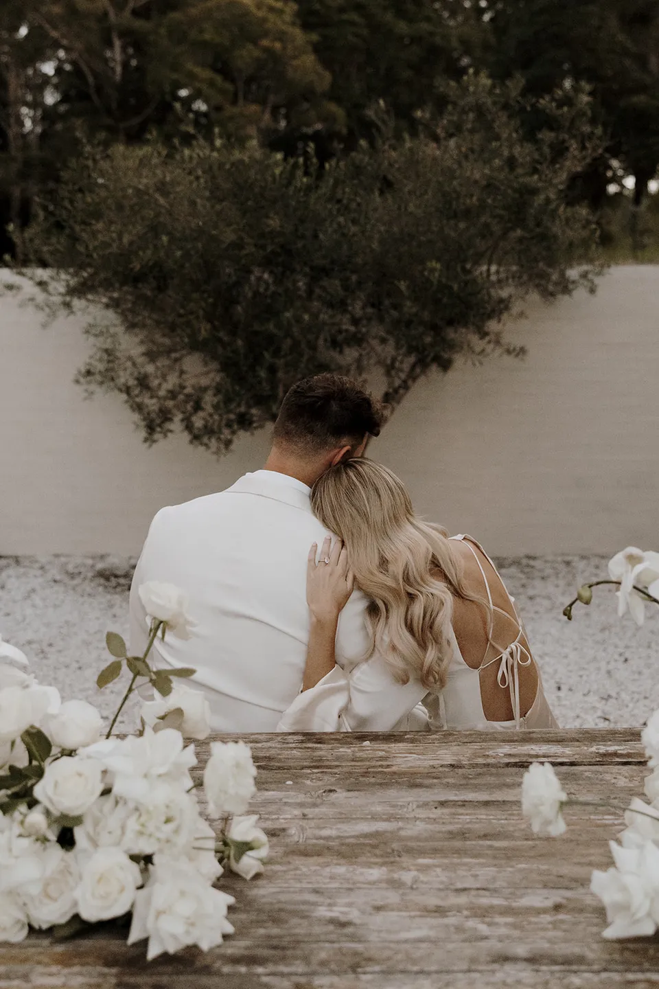 Gabrielle resting her head against Greg at the courtyard table, with white flowers in the foreground at Roundhill Retreat in Obi Obi, Sunshine Coast Hinterland, for their Elopement Collective elopement.