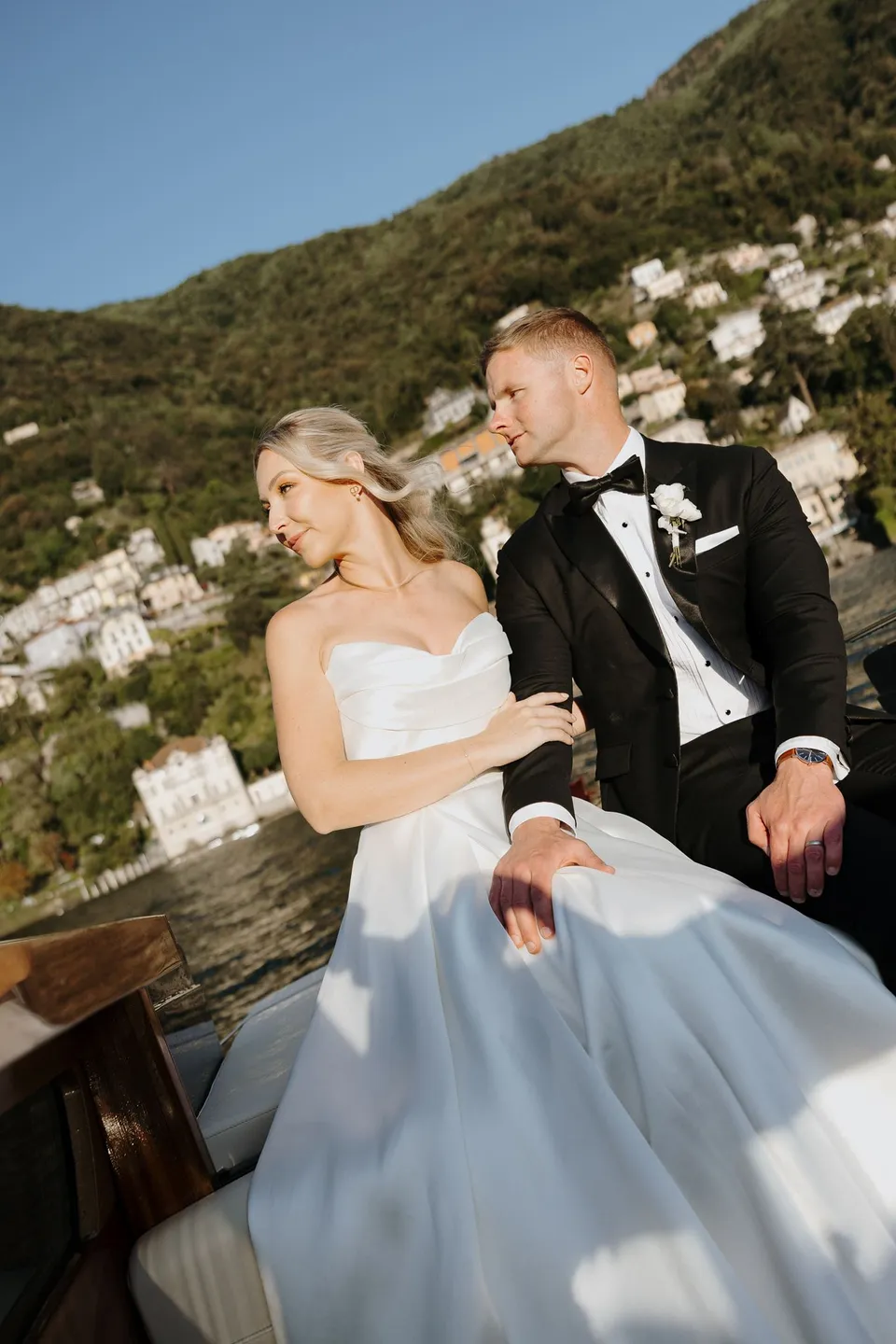 Zoe and Charlton standing together beside the wooden boat rail with a hillside village across the water, on Lake Como in Lombardy, Italy, for an Elopement Collective elopement. Photo by George Bowden.