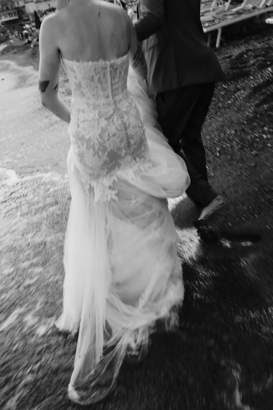 Black and white close view of Skyler’s lace dress and trailing skirt as she and Zach step together through shallow water, in Positano on the Amalfi Coast, Italy, for an Elopement Collective elopement. Photograph by Jason Corroto.