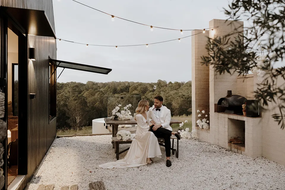 Gabrielle and Greg sitting together in the cabin courtyard beneath festoon lights, with the outdoor fireplace beside them at Roundhill Retreat in Obi Obi, Sunshine Coast Hinterland, for their Elopement Collective elopement.
