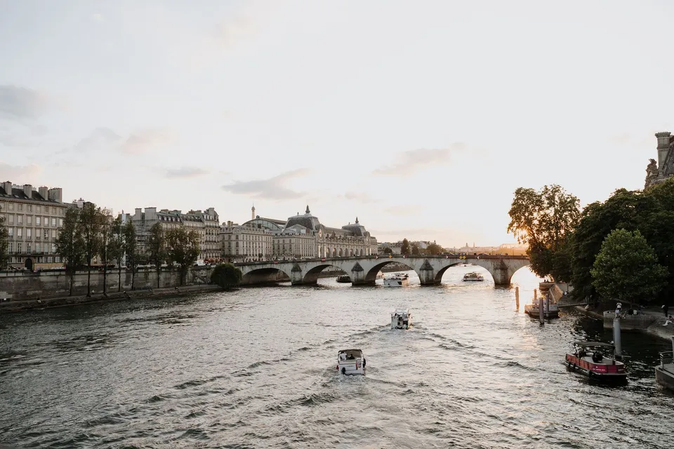 A boat travelling along sunlit water on the Seine towards an arched bridge, with buildings and trees along the banks, beside the Seine in Paris, France, during an Elopement Collective elopement. Photograph by George Bowden.