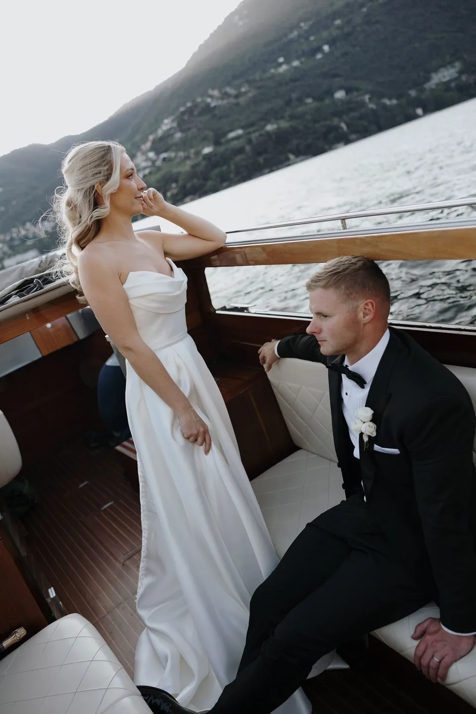 Zoe standing beside the boat window while Charlton sits on a white bench, with the lake and wooded hills beyond, on Lake Como in Lombardy, Italy, for an Elopement Collective elopement. Photo by George Bowden.
