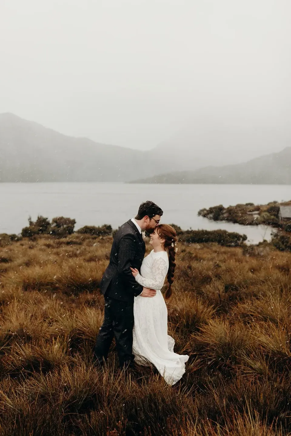 A couple kiss in golden grass beside a misty mountain lake.