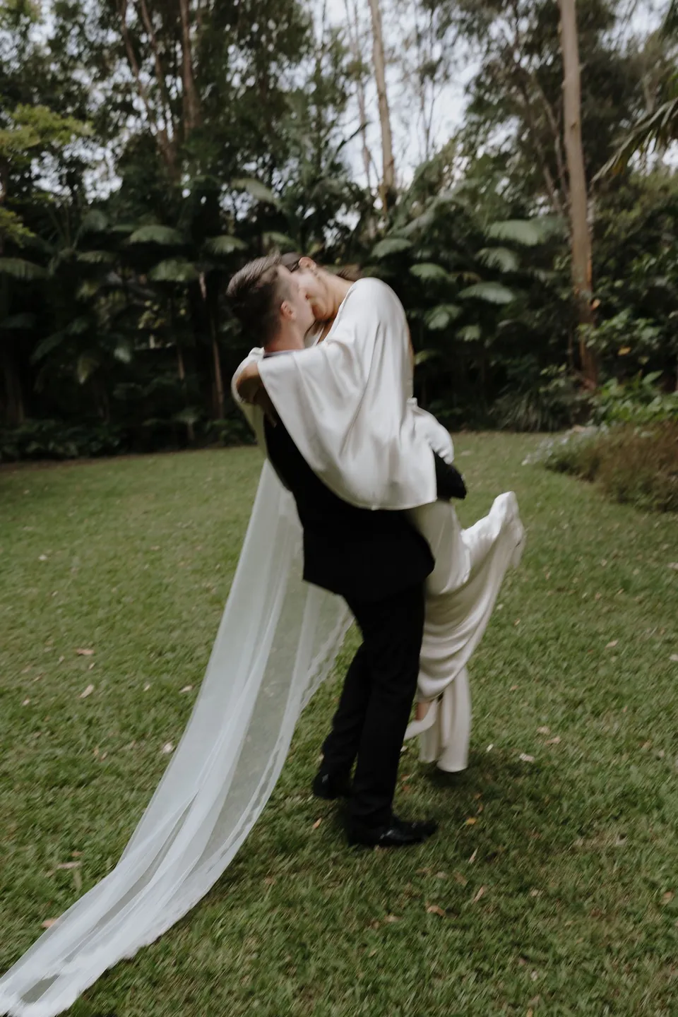 Laura and Matthew kissing as he lifts her from the grass. At Fig Tree Villa in Ewingsdale, Byron Bay, for an Elopement Collective elopement. Photograph by George Bowden.