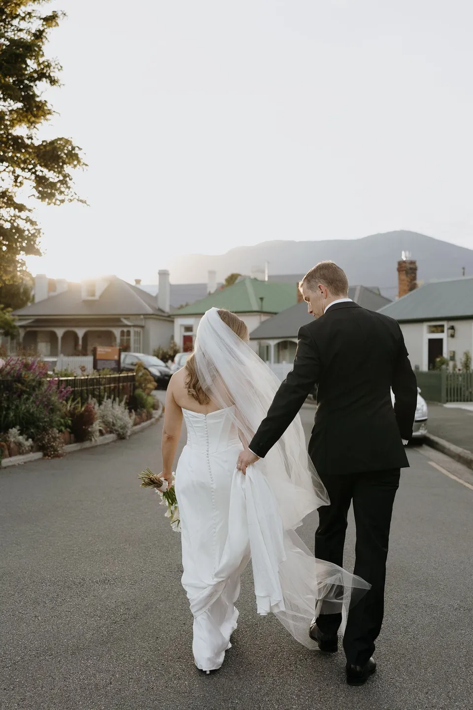 Tanesha and Andrew walking away hand in hand along the curved cottage-lined street, her veil catching the evening light, at Arthur Circus in Battery Point, Hobart, Tasmania, Australia, for an Elopement Collective summer elopement. Photograph by George Bowden.
