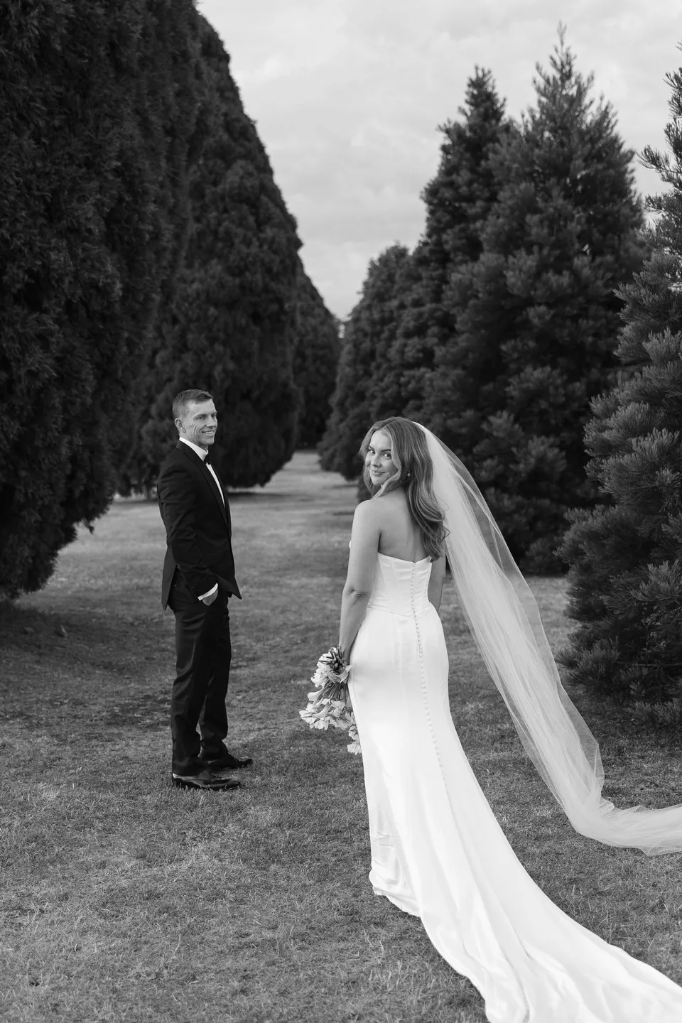 Black and white portrait of Tanesha looking back while Andrew stands beside her on a path framed by tall evergreen trees, at Hobart, Tasmania, Australia, for an Elopement Collective summer elopement. Photograph by George Bowden.