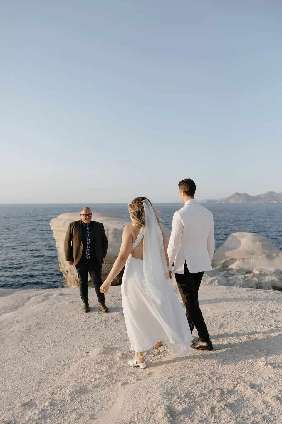 Georgina and Brad holding hands and walking towards celebrant Josh Withers, who is waiting on the rocks above the sea, at Sarakiniko on Milos in the Cyclades, Greece, during an Elopement Collective elopement. Photograph by George Bowden.
