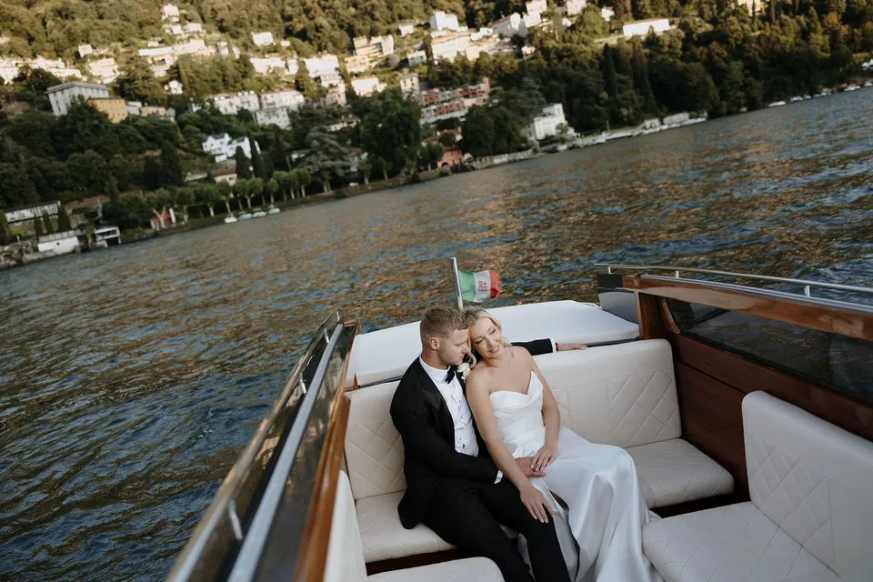 Zoe and Charlton kissing on a white boat seat beneath an Italian flag, with a hillside village across the water, on Lake Como in Lombardy, Italy, for an Elopement Collective elopement. Photo by George Bowden.