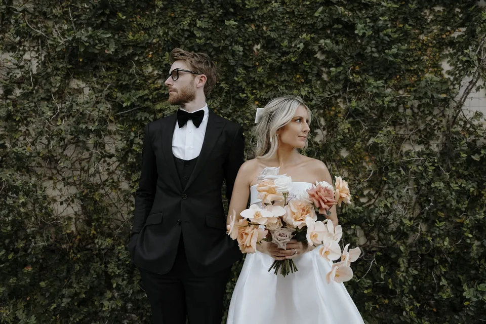 Danielle holding her peach and white bouquet beside Adam in front of a leafy ivy wall, during Danielle and Adam’s Elopement Collective elopement at The Calile Hotel in Fortitude Valley, Brisbane.