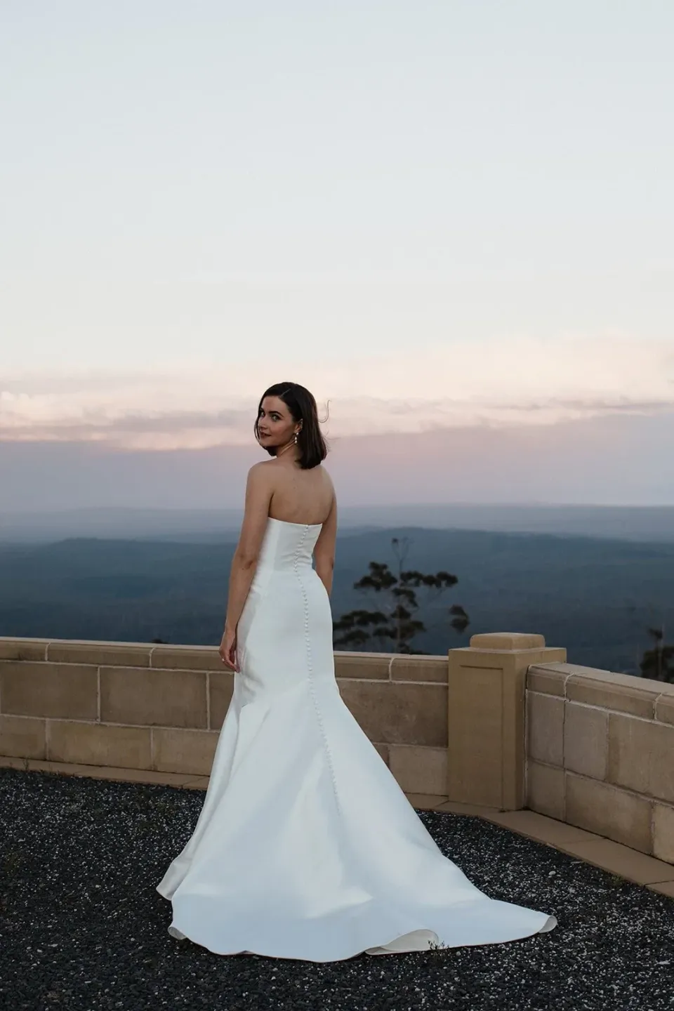 A bride looks back from a low stone wall overlooking hills at dusk.