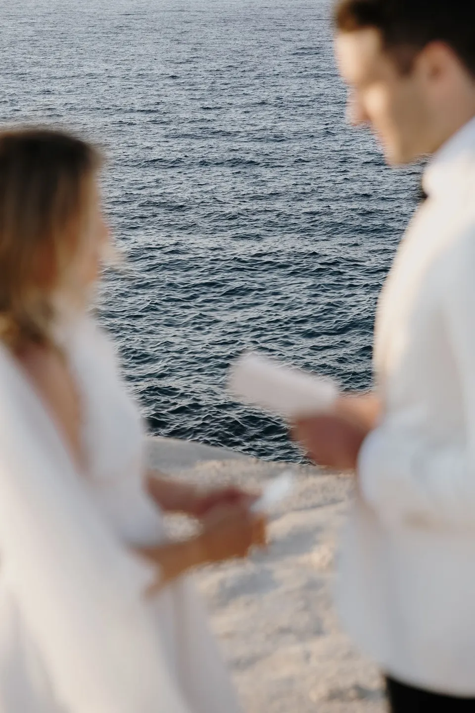A soft-focus view of Brad holding his vow book and Georgina’s hand as she faces him, with seawater behind them, at Sarakiniko on Milos in the Cyclades, Greece, during an Elopement Collective elopement. Photograph by George Bowden.