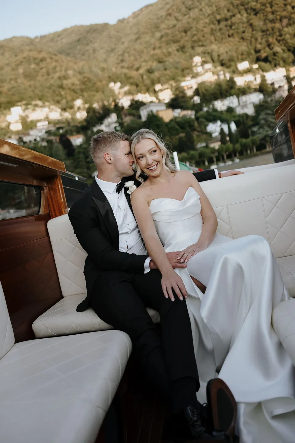 Charlton leaning close to Zoe as they sit together inside the wooden boat, with the lake visible through the open window, on Lake Como in Lombardy, Italy, for an Elopement Collective elopement. Photo by George Bowden.
