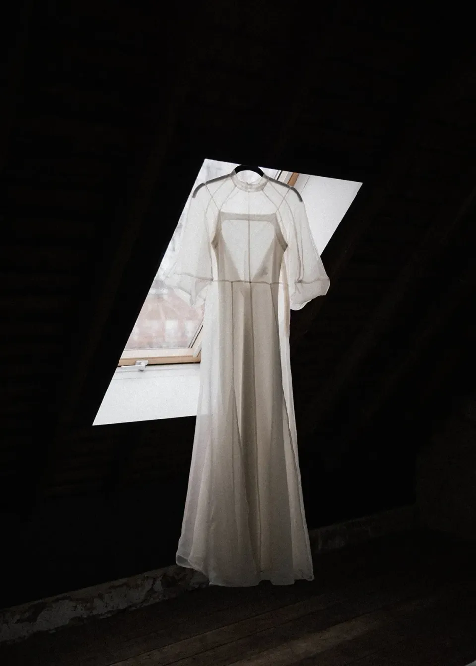 A wedding dress hangs in the light from a small attic window.