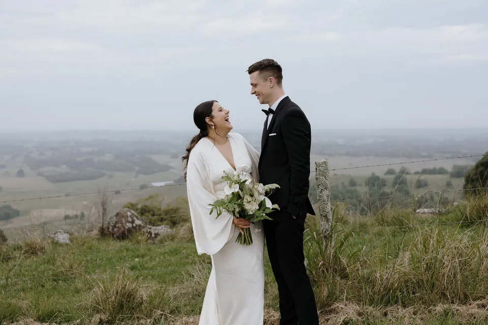 Laura and Matthew laughing together at a lookout above the green countryside. In the Byron Bay region of northern New South Wales, for an Elopement Collective elopement. Photograph by George Bowden.