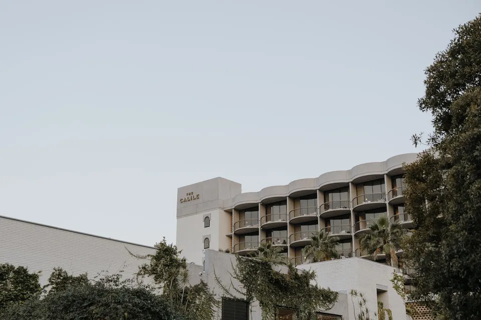 The Calile Hotel’s curved balconies and signed white brick exterior above leafy trees, during Danielle and Adam’s Elopement Collective elopement at The Calile Hotel in Fortitude Valley, Brisbane.