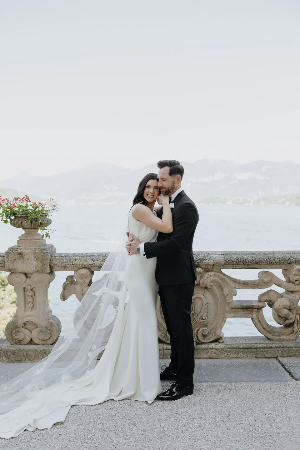 Lyndal and Lachlan embracing beside a stone balustrade and flower-filled urn at Villa del Balbianello, with the lake and mountains behind them, at Lake Como in Lombardy, Italy, for an Elopement Collective elopement. Photograph by George Bowden.