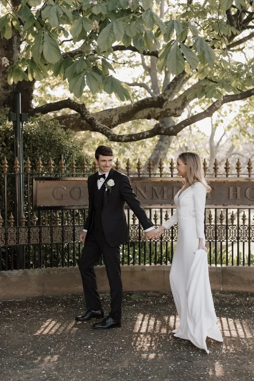 A couple hold hands beneath a spreading tree beside an iron garden fence.