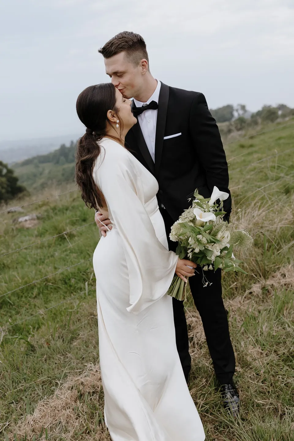 Laura and Matthew kissing with a broad hinterland view behind them. In the Byron Bay region of northern New South Wales, for an Elopement Collective elopement. Photograph by George Bowden.