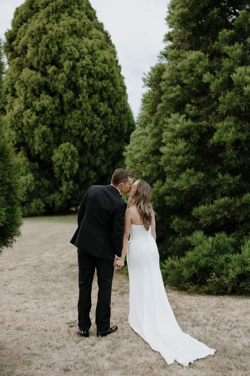 Tanesha and Andrew kissing while holding hands on a pale path between tall evergreen trees, at Hobart, Tasmania, Australia, for an Elopement Collective summer elopement. Photograph by George Bowden.