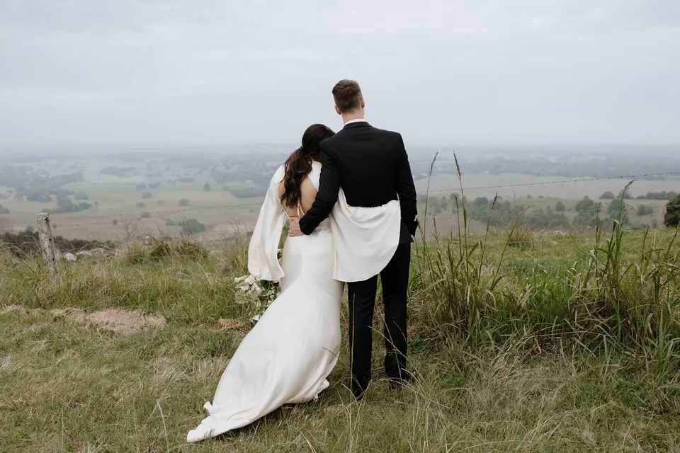 Laura and Matthew looking out over the countryside with their arms around each other. In the Byron Bay region of northern New South Wales, for an Elopement Collective elopement. Photograph by George Bowden.