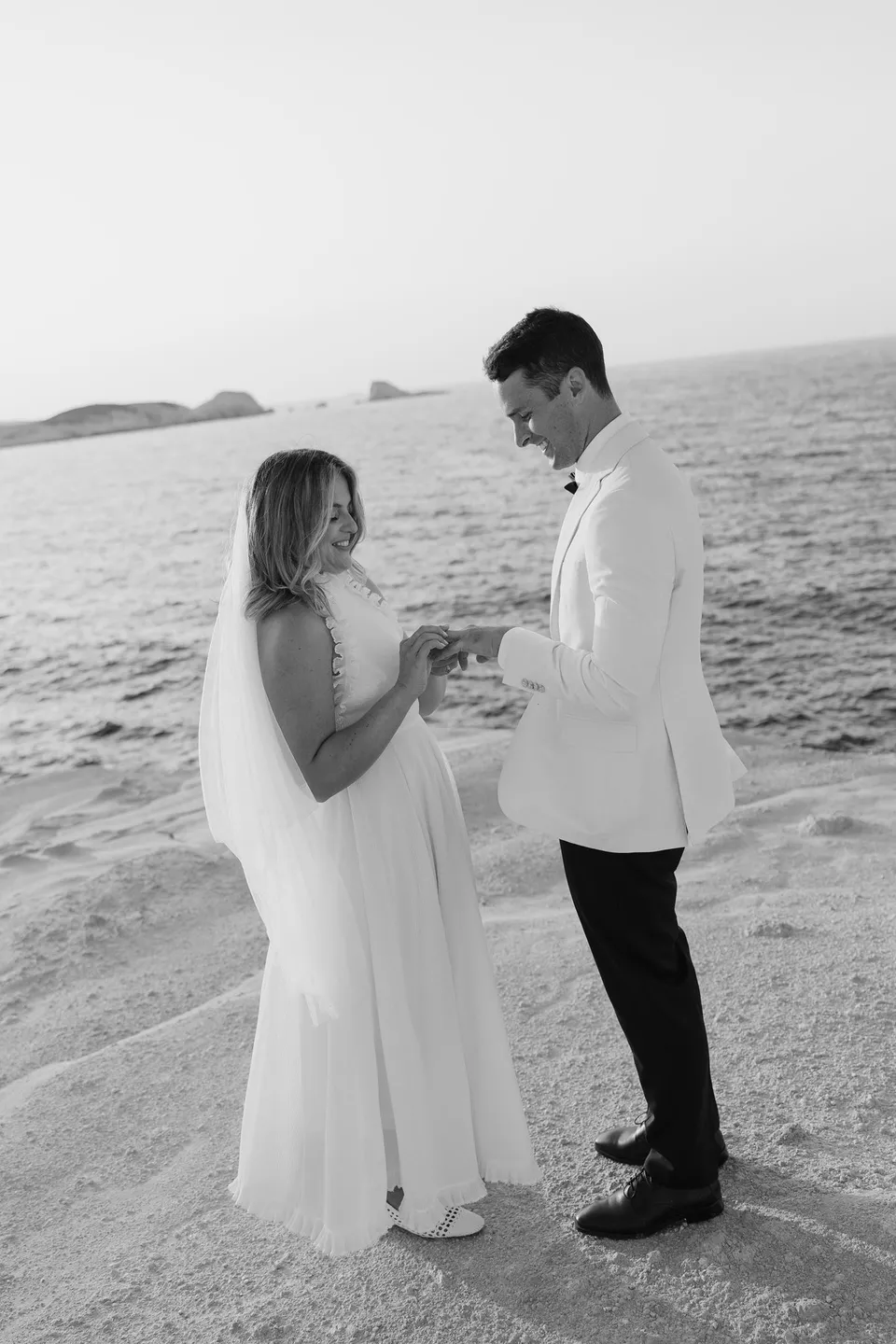 Black and white photograph of Georgina smiling as she places a wedding ring on Brad’s finger beside the sea, at Sarakiniko on Milos in the Cyclades, Greece, during an Elopement Collective elopement. Photograph by George Bowden.