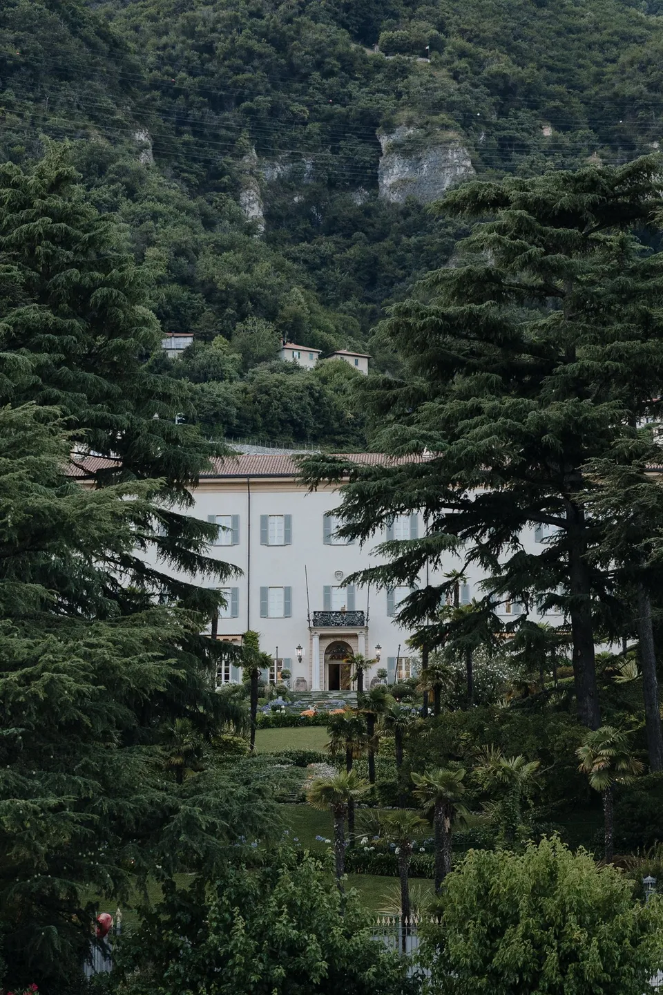 Passalacqua’s pale villa and shuttered windows seen through tall trees and layered garden greenery, at Passalacqua in Moltrasio, Lake Como, Italy, for an Elopement Collective elopement. Photo by George Bowden.