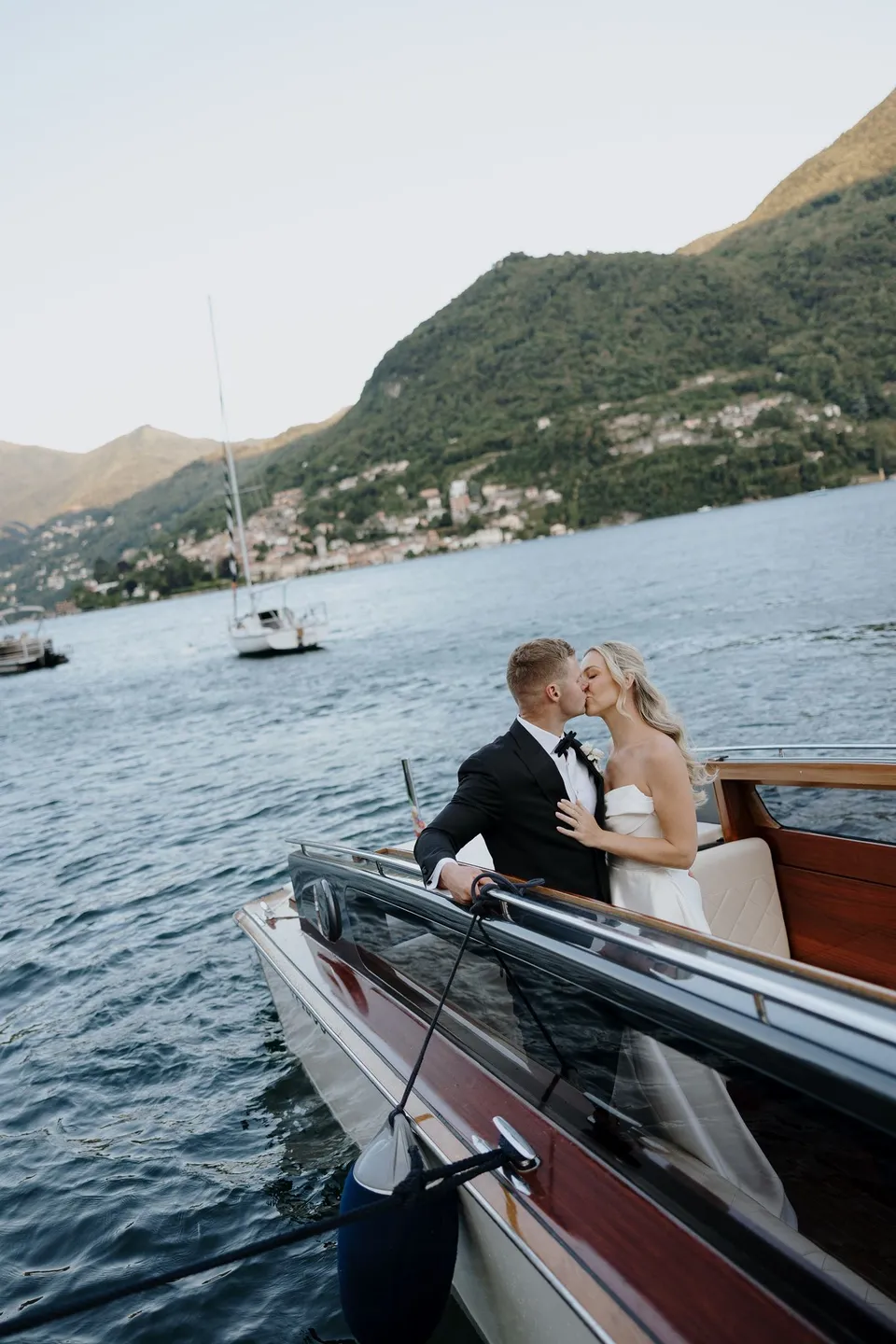 Zoe and Charlton kissing beside the polished wooden edge of a boat, with open water, a sailboat and mountains behind them, on Lake Como in Lombardy, Italy, for an Elopement Collective elopement. Photo by George Bowden.