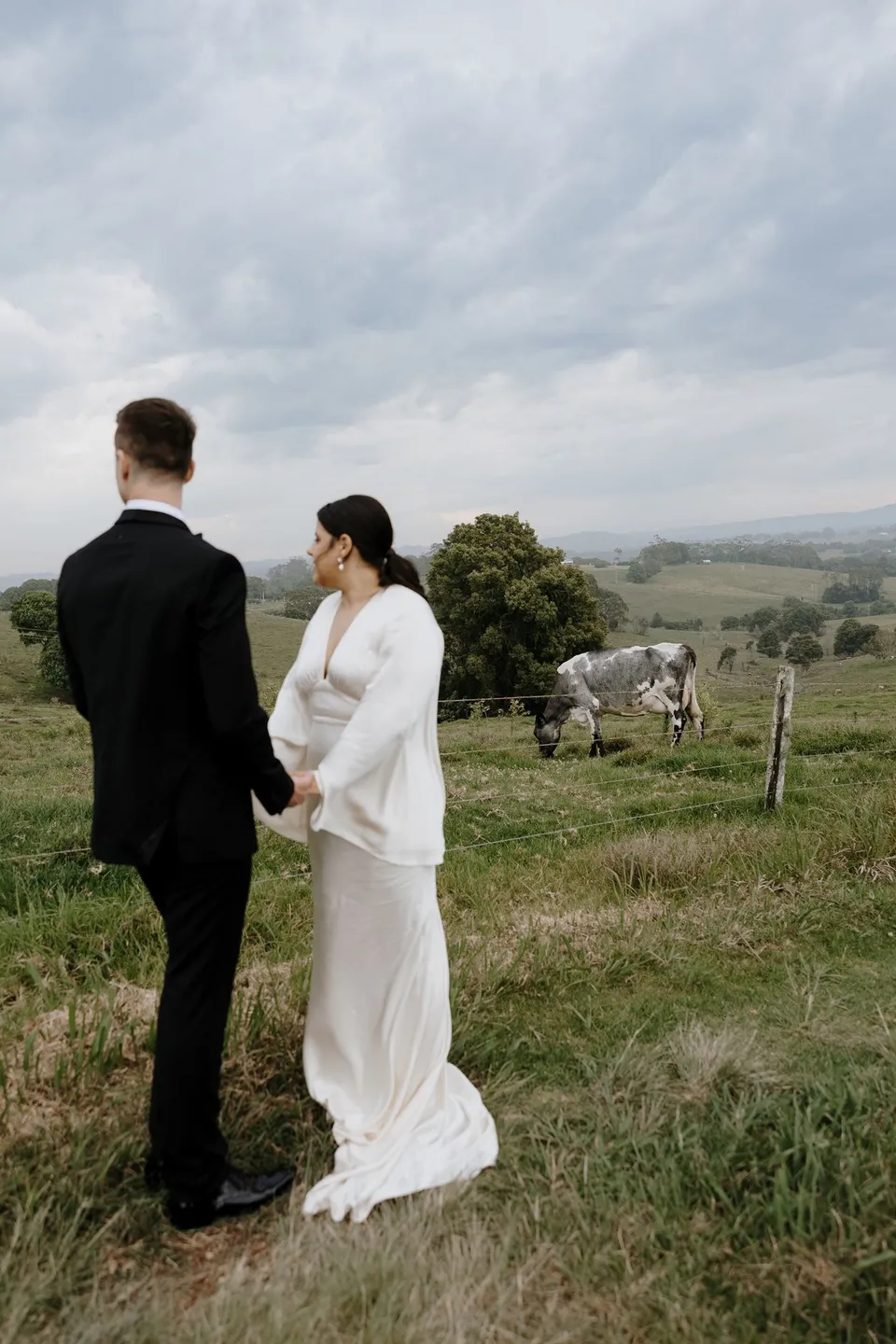 Laura and Matthew holding hands in a paddock beneath a cloud-filled sky. In the Byron Bay region of northern New South Wales, for an Elopement Collective elopement. Photograph by George Bowden.