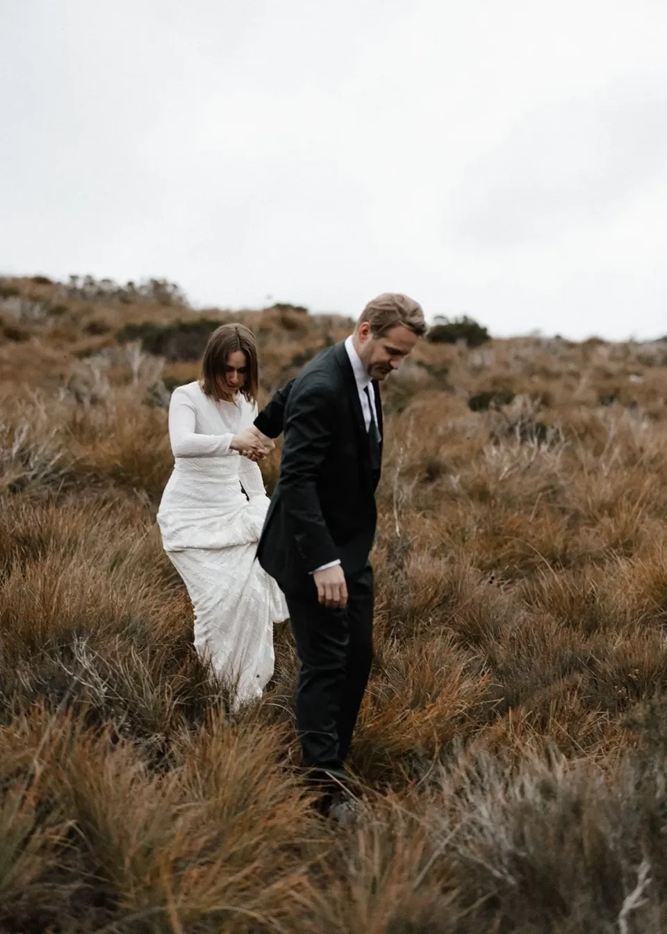 A couple look down as they walk together through long grass.