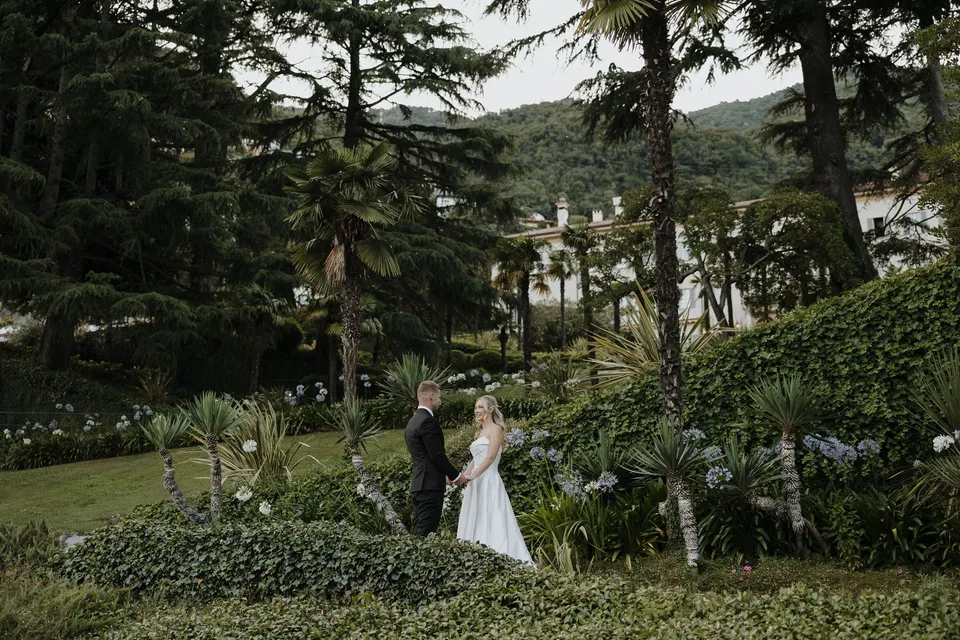 Zoe and Charlton standing together on a path among terraced hedges and tall trees, with a glimpse of the lake beyond, at Passalacqua in Moltrasio, Lake Como, Italy, for an Elopement Collective elopement. Photo by George Bowden.