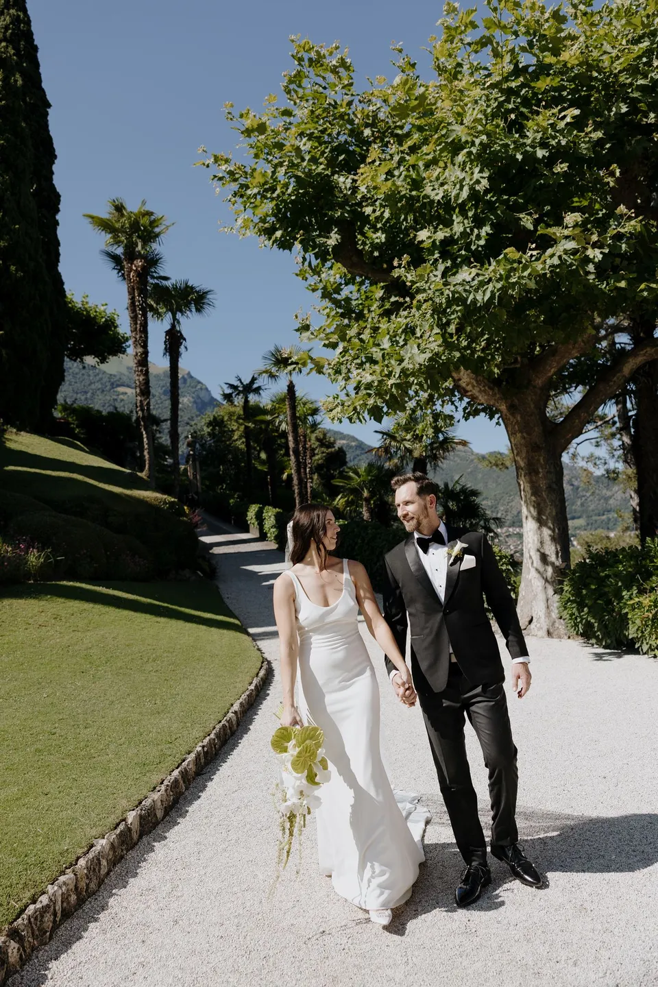 Lyndal and Lachlan walking along a garden path and steps at Villa del Balbianello, carrying a white bouquet by Garden of Love, at Lake Como in Lombardy, Italy, for an Elopement Collective elopement. Photograph by George Bowden.