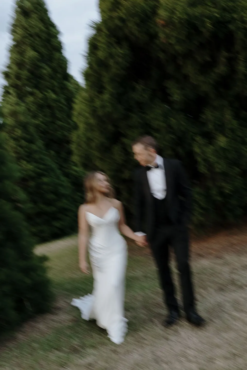 Softly blurred photograph of Tanesha and Andrew walking hand in hand along a tree-lined path, at Hobart, Tasmania, Australia, for an Elopement Collective summer elopement. Photograph by George Bowden.