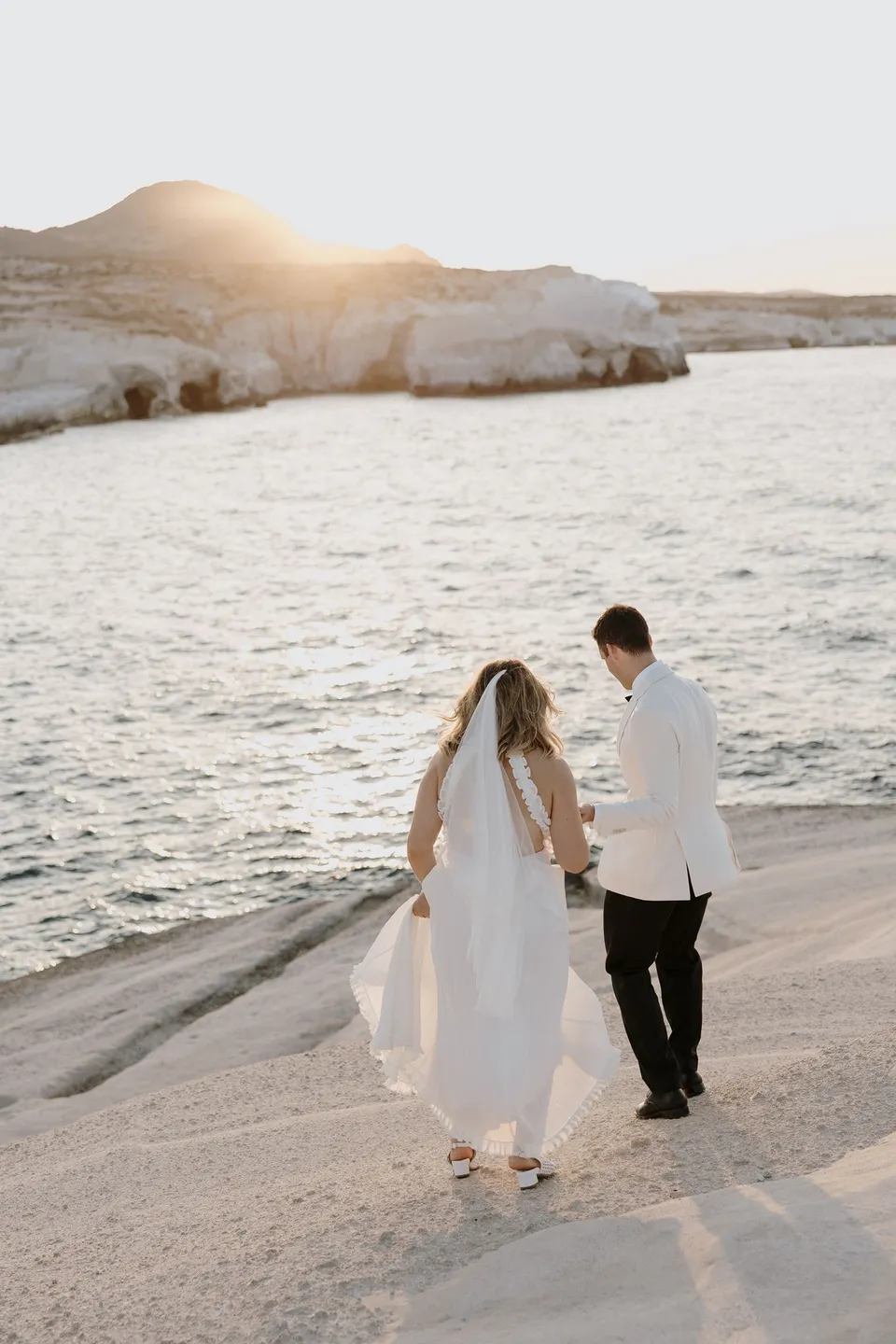 Georgina and Brad seen from behind holding hands as they walk beside an inlet in warm evening light, at Sarakiniko on Milos in the Cyclades, Greece, during an Elopement Collective elopement. Photograph by George Bowden.