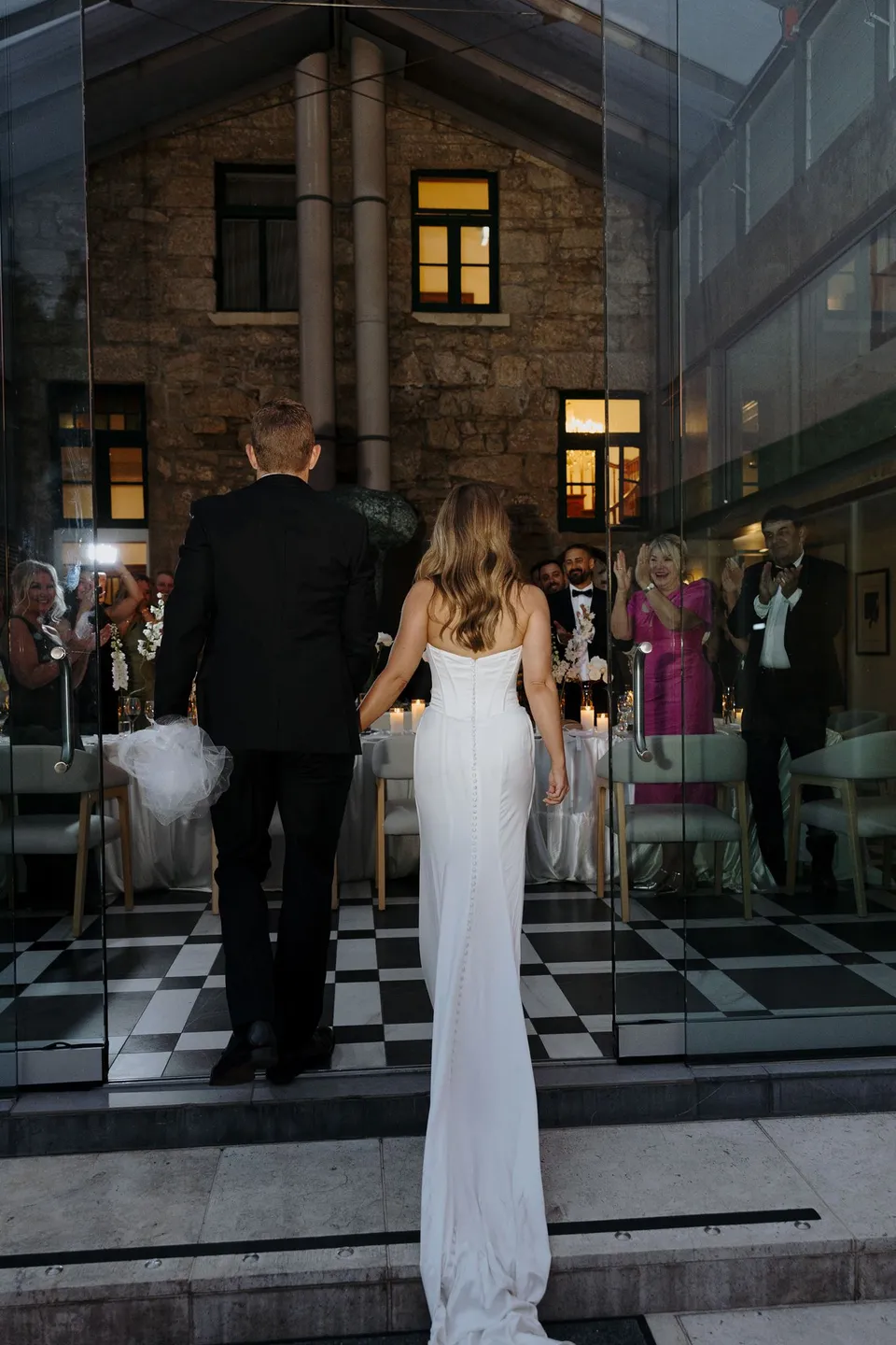 Tanesha and Andrew entering the glass conservatory together across a black and white tiled floor as their guests look on, at Islington Hotel in South Hobart, Tasmania, Australia, for an Elopement Collective summer elopement. Photograph by George Bowden.
