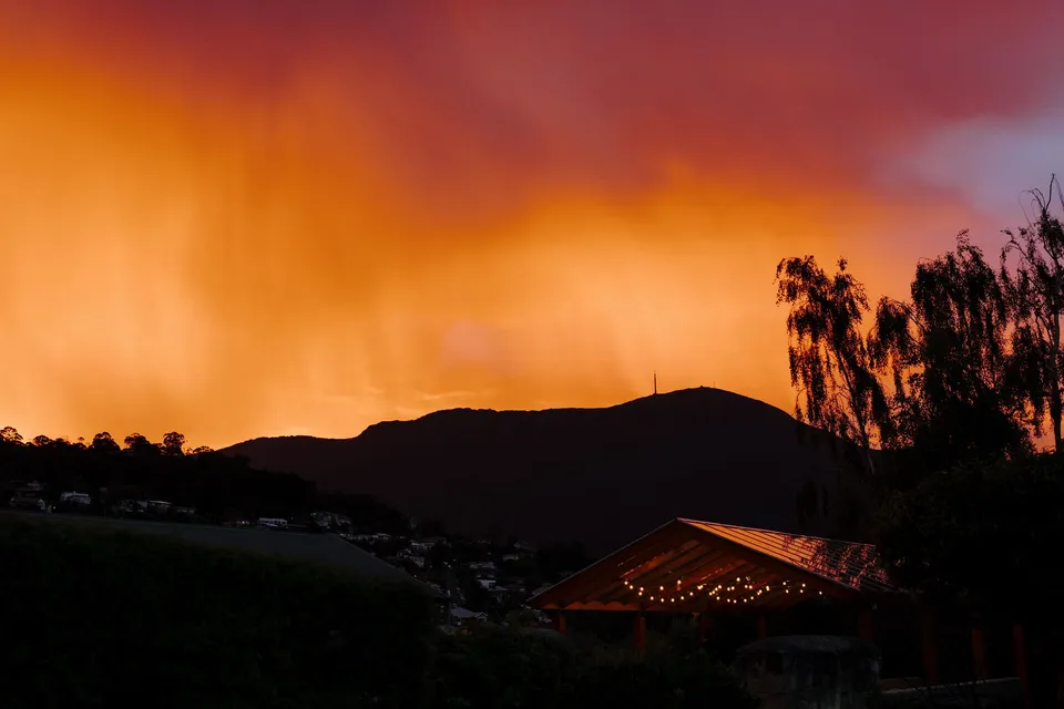A vivid orange and pink summer sunset above the silhouetted mountain, with hotel roofs and trees in the foreground, at Islington Hotel in South Hobart, Tasmania, Australia, for an Elopement Collective summer elopement. Photograph by George Bowden.