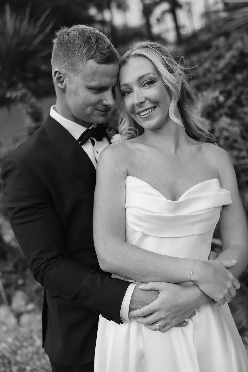 Black and white portrait of Charlton embracing Zoe from behind as she smiles towards the camera, at Passalacqua in Moltrasio, Lake Como, Italy, for an Elopement Collective elopement. Photo by George Bowden.