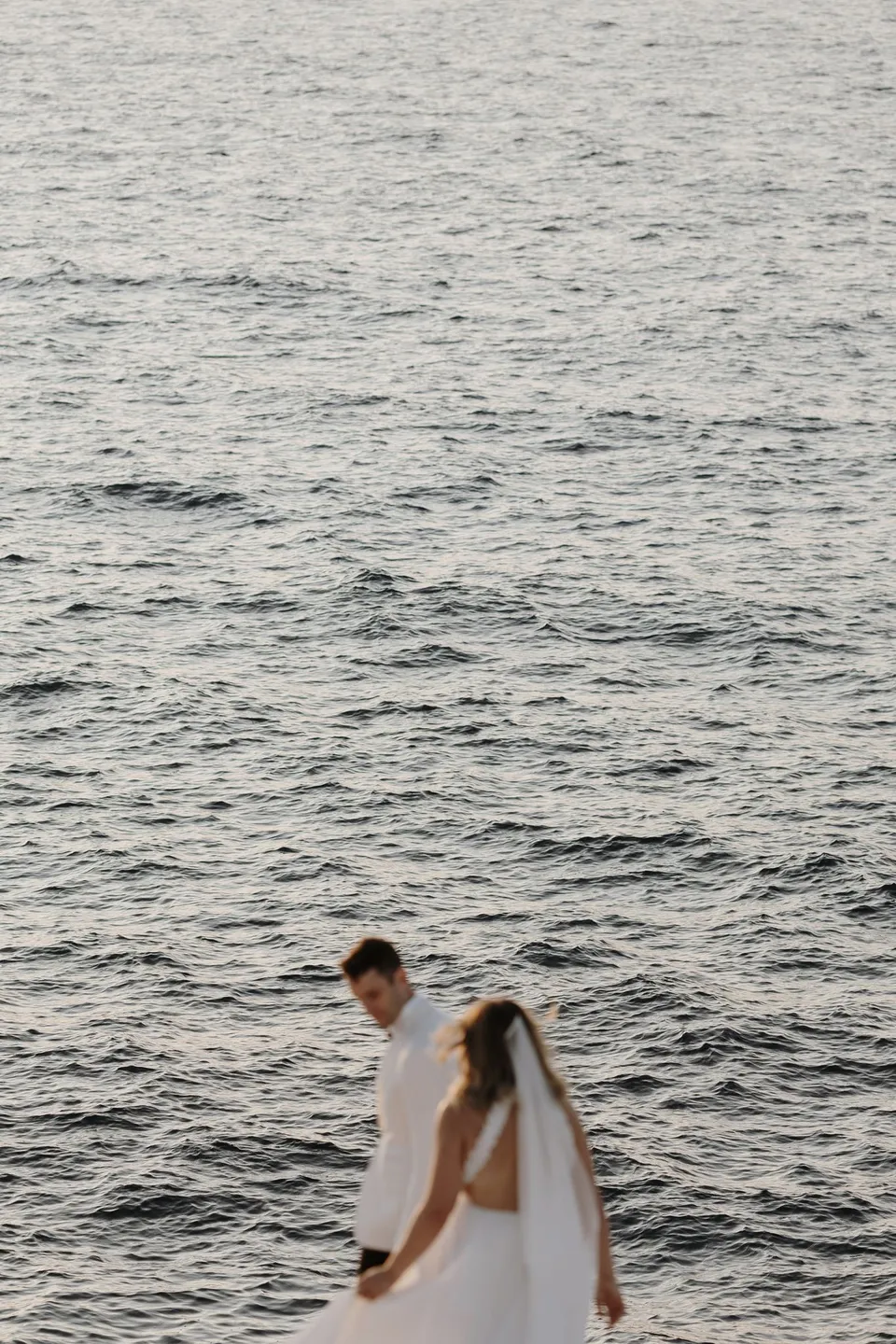 Georgina holding Brad’s arm beside the inlet, with rippling blue seawater filling the background, at Sarakiniko on Milos in the Cyclades, Greece, during an Elopement Collective elopement. Photograph by George Bowden.