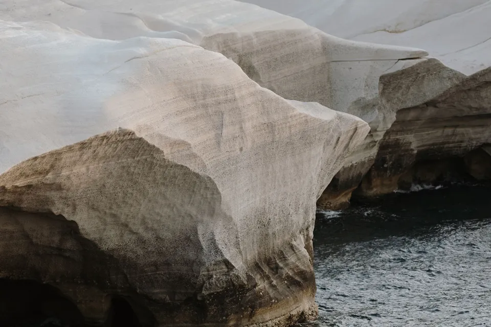 Close view of layered, weathered white rocks forming an arch above blue water, at Sarakiniko on Milos in the Cyclades, Greece, during an Elopement Collective elopement. Photograph by George Bowden.