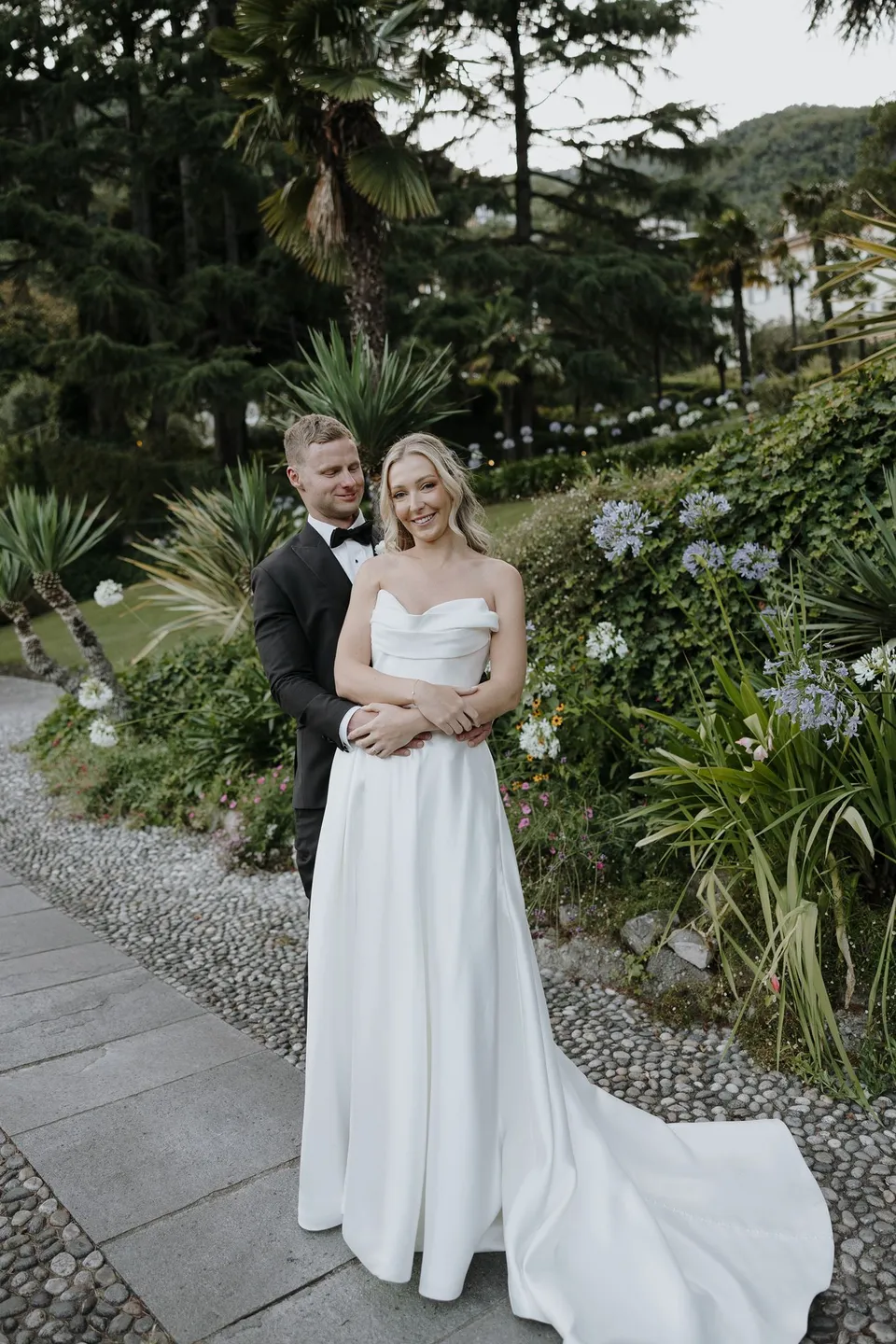 Full-length portrait of Charlton embracing Zoe from behind on a garden path, with her white train spread out beside the greenery, at Passalacqua in Moltrasio, Lake Como, Italy, for an Elopement Collective elopement. Photo by George Bowden.