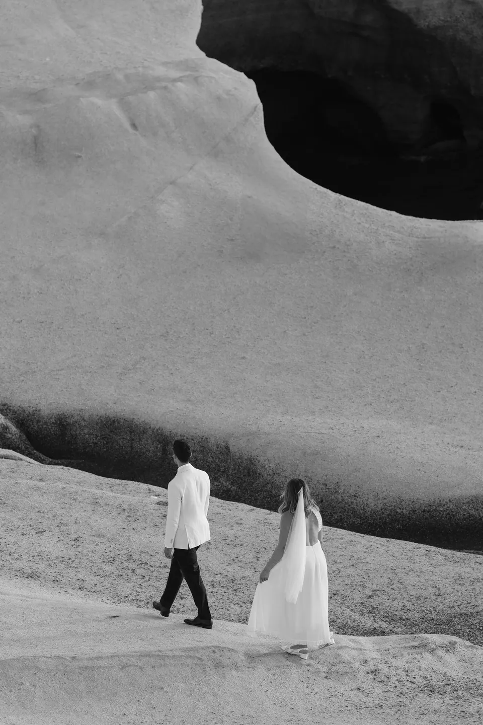 Black and white photograph of Georgina and Brad walking across pale rocks below a large dark opening in the cliff, at Sarakiniko on Milos in the Cyclades, Greece, during an Elopement Collective elopement. Photograph by George Bowden.