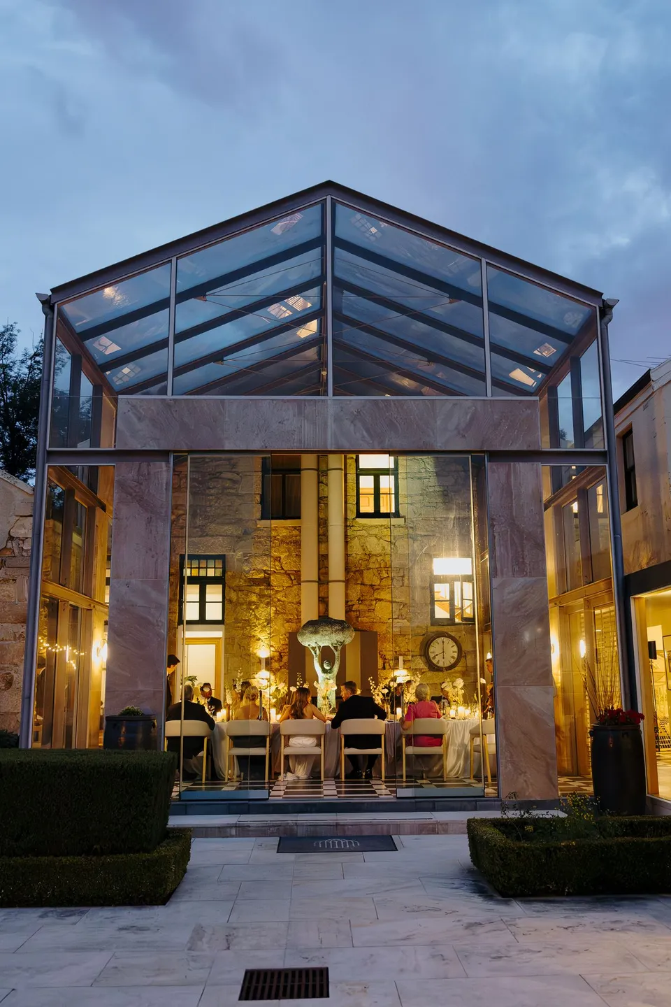 The illuminated glass conservatory seen from the garden at dusk, with reception guests seated inside, at Islington Hotel in South Hobart, Tasmania, Australia, for an Elopement Collective summer elopement. Photograph by George Bowden.