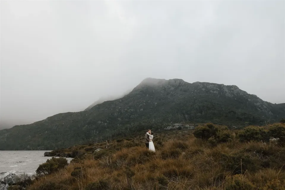 A bride stands in alpine scrub between a lake and a mist-covered mountain.