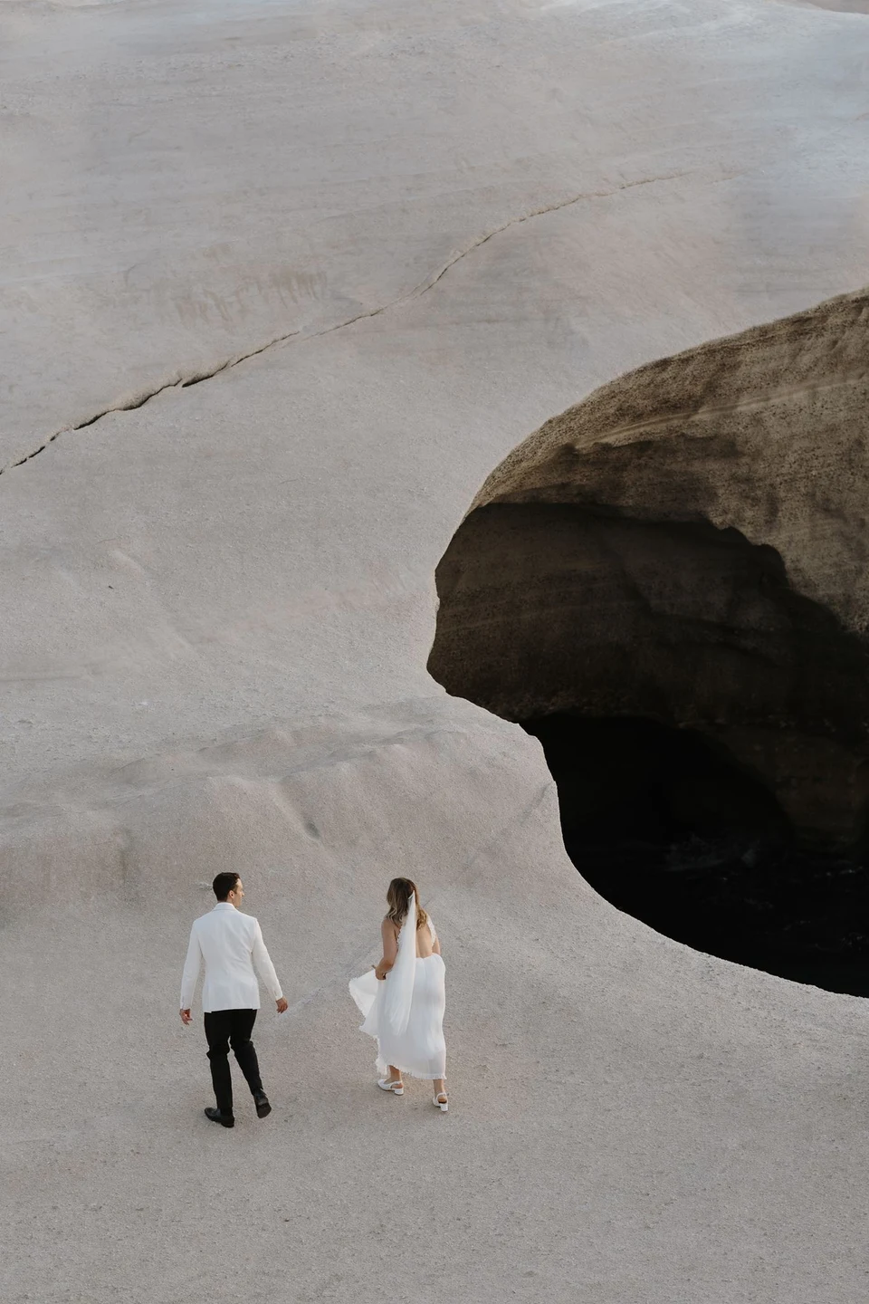 Georgina and Brad seen from behind walking down a smooth pale rock slope towards an inlet, at Sarakiniko on Milos in the Cyclades, Greece, during an Elopement Collective elopement. Photograph by George Bowden.