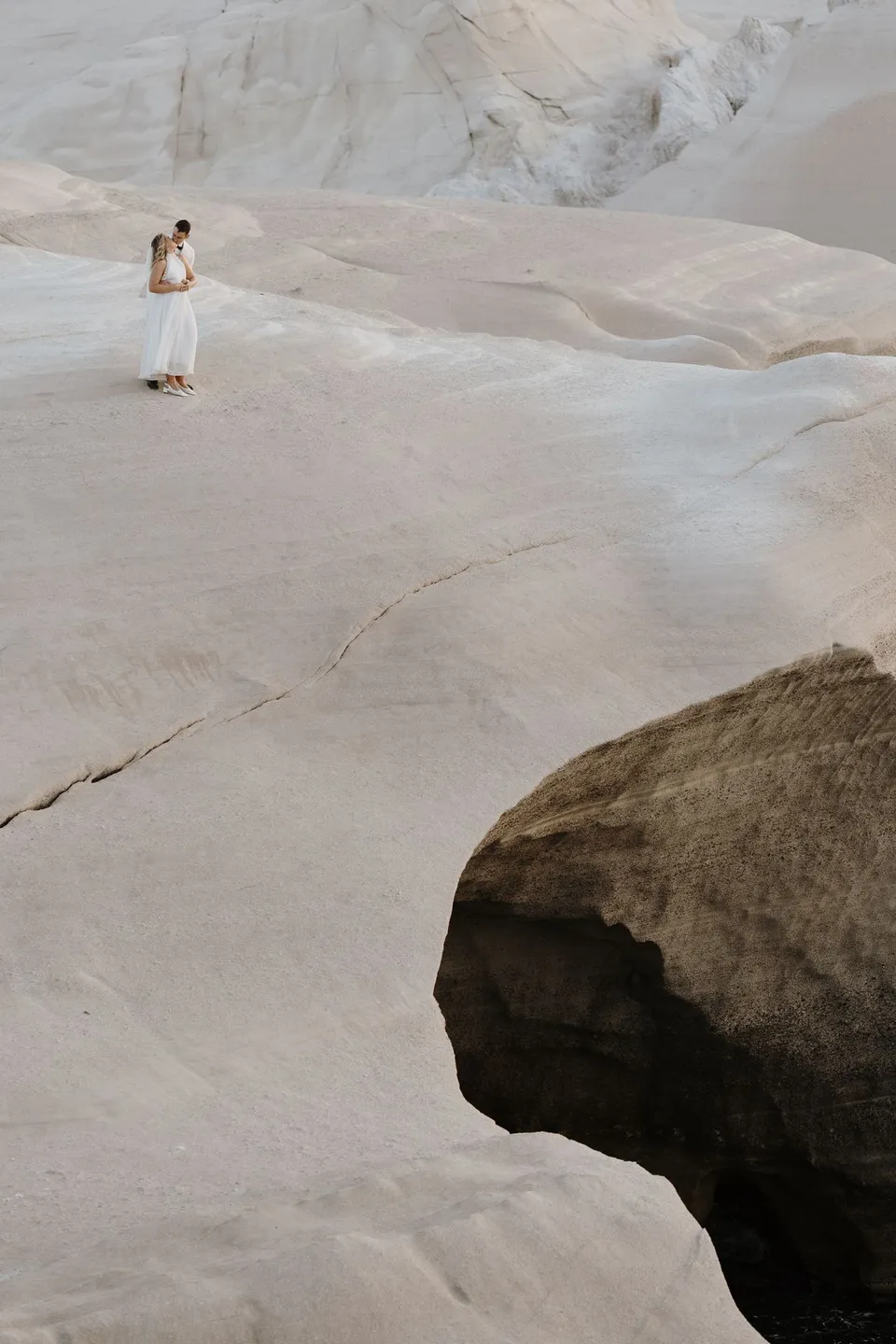 Georgina and Brad embracing on a broad smooth white rock plateau beside a dark inlet, at Sarakiniko on Milos in the Cyclades, Greece, during an Elopement Collective elopement. Photograph by George Bowden.