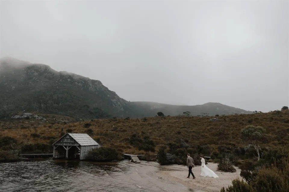 A couple walk along a mountain lakeshore beside a small timber boatshed.