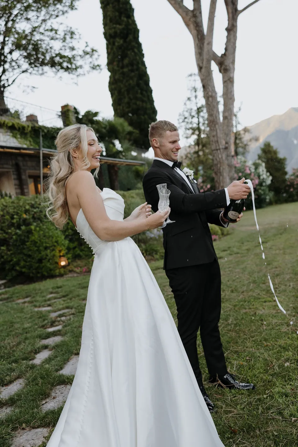 Charlton spraying champagne beside Zoe on the lakeside lawn as she holds a flute and watches, at Passalacqua in Moltrasio, Lake Como, Italy, for an Elopement Collective elopement. Photo by George Bowden.