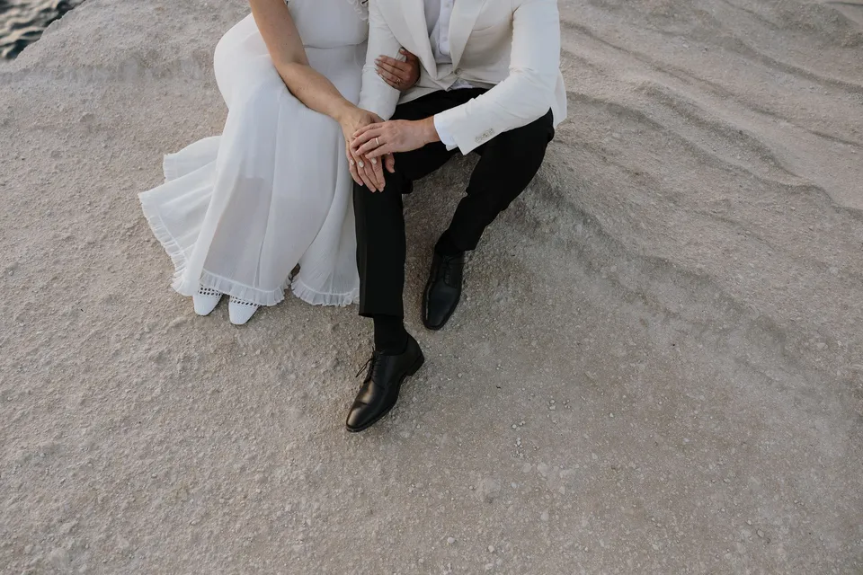 Close view from above of Georgina and Brad’s hands and wedding rings as they sit together, with her dress, his trousers and woven shoes visible, at Sarakiniko on Milos in the Cyclades, Greece, during an Elopement Collective elopement. Photograph by George Bowden.